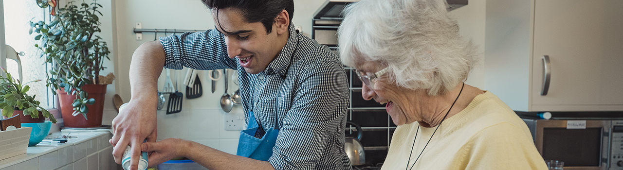 Teenage boy making fruit compote with his grandmother in the kitchen of her home. He is adding squirty cream to the bowls.